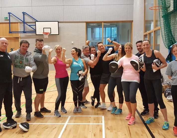 Group of Glasgow Kelvin College staff and students smiling and flexing after a fitness session in the gym hall, wearing boxing gloves and pads. Group of Glasgow Kelvin College staff and students smiling and flexing after a fitness session in the gym hall, wearing boxing gloves and pads.